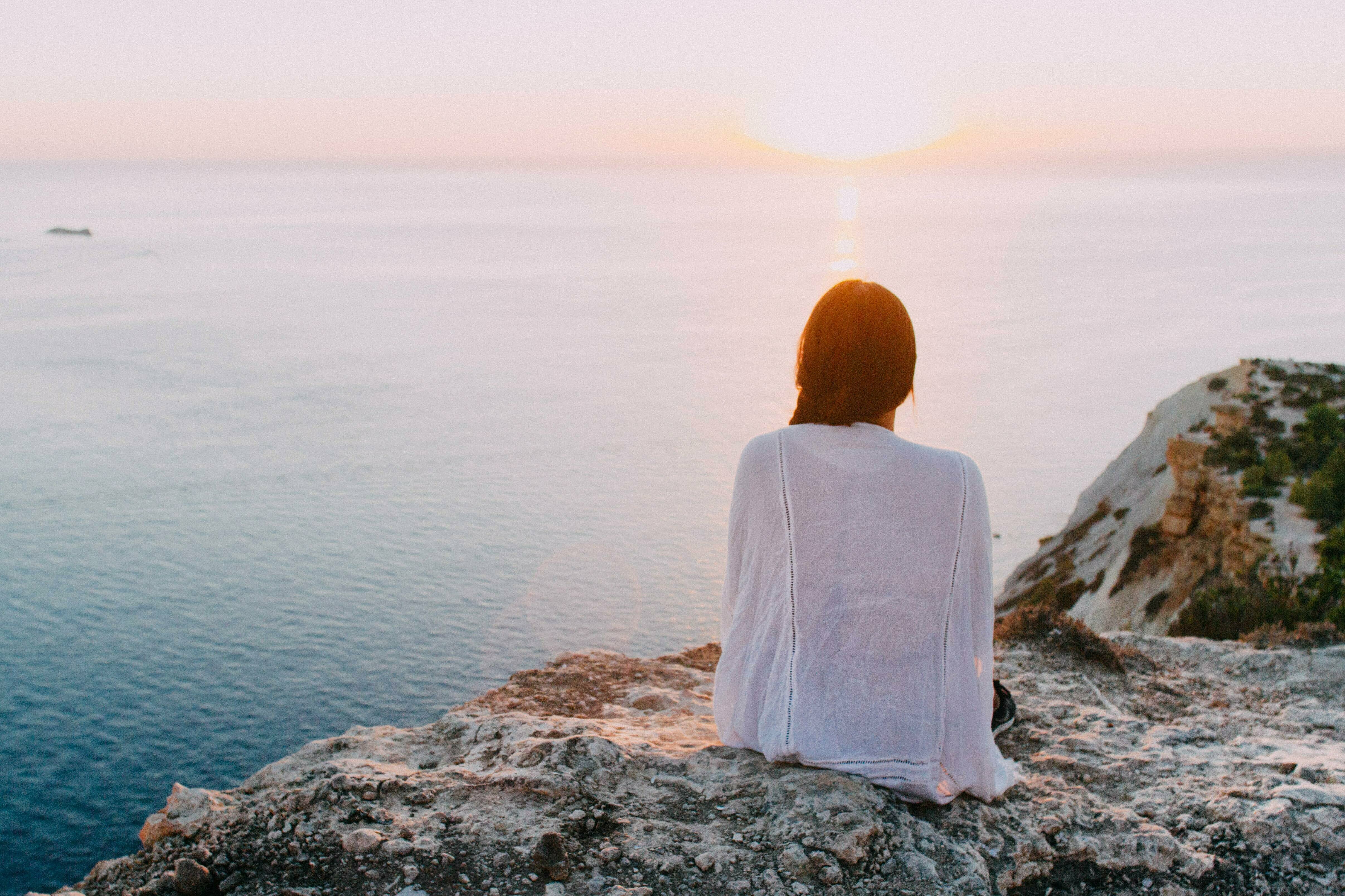 Frau blickt, sitzend auf einem Fels, auf den Sonnenaufgang über dem Meer